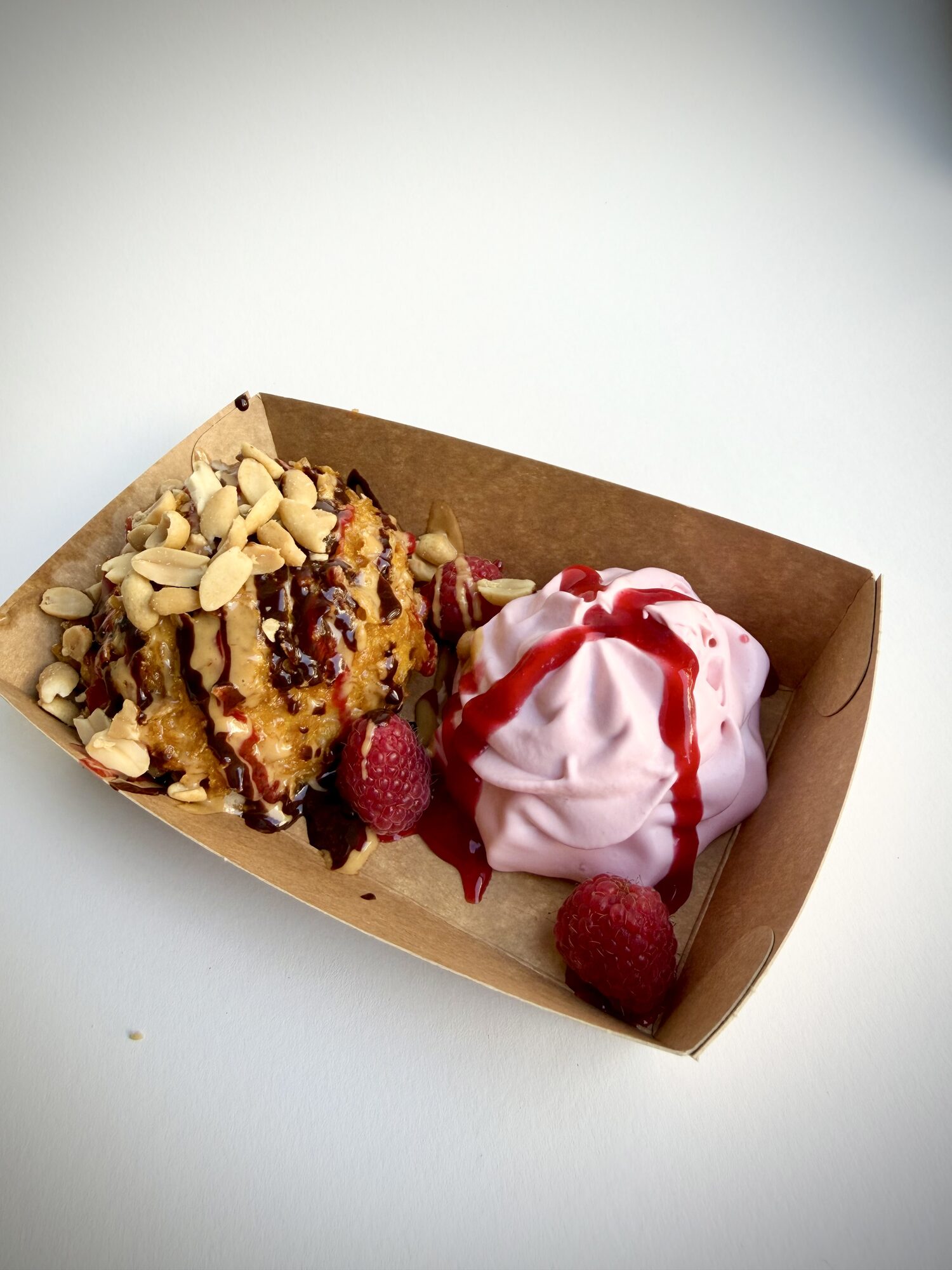 A fried scoop of banana ice cream with roasted peanuts and chocolate sauce, next to a pink raspberry-lime cream with raspberry sauce and fresh raspberries, in a kraft paper tray.