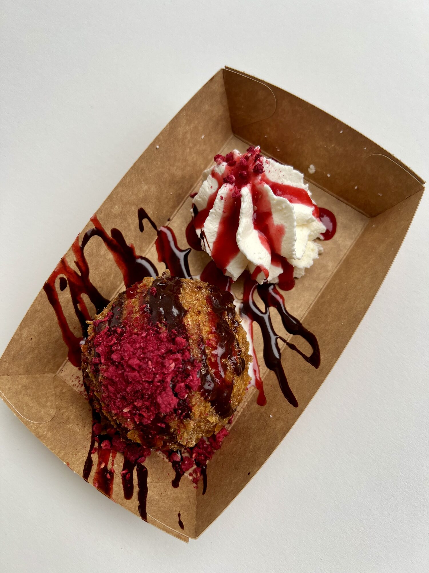 A fried scoop of vanilla ice cream with raspberry sauce, raspberry dust and whipped cream in a kraft paper tray.